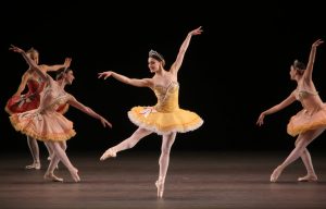 Ballerina on her toes in a yellow tutu surrounded by srps de ballet dancers