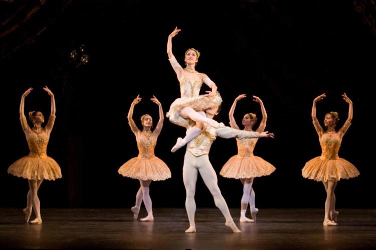 Ballerina sits on shoulder of her partner and four corps de ballet dancers surround them. All ballerinas are wearing light pink tutus, male dancer is in white.